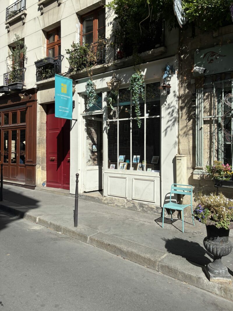 Façade de la galerie La Capitainerie de l'Ile à Paris, vitrine ornée de plantes suspendues et de tirages de Sophie Moisan, chaise bleue sur le trottoir