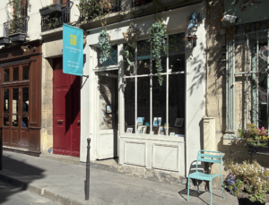 Façade de la galerie La Capitainerie de l'Ile à Paris, vitrine ornée de plantes suspendues et de tirages de Sophie Moisan, chaise bleue sur le trottoir