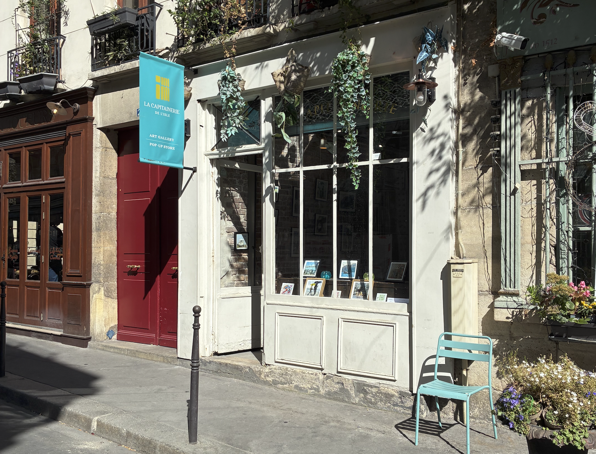 Façade de la galerie La Capitainerie de l'Ile à Paris, vitrine ornée de plantes suspendues et de tirages de Sophie Moisan, chaise bleue sur le trottoir