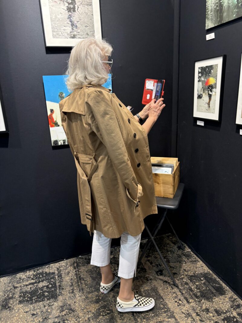 Visiteuse en trench beige photographiant avec son téléphone les photographies de pluie de Sophie Moisan sur le stand Artiti.art